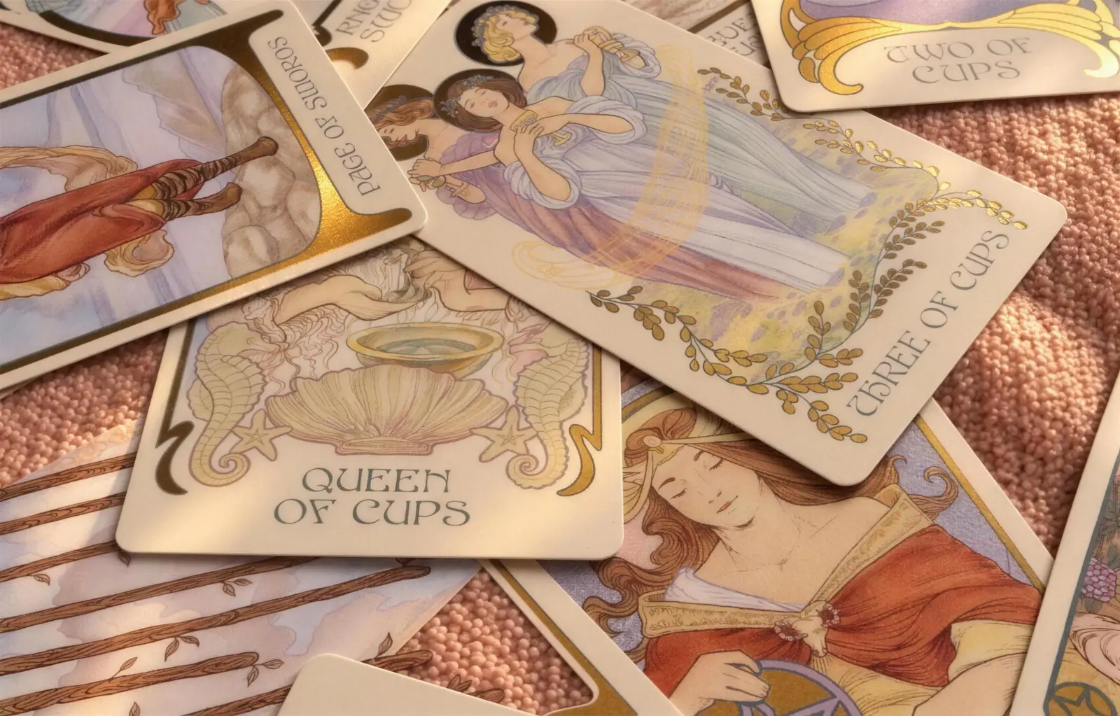 Cartas de tarô espalhadas sobre um tecido rosa, com a Rainha de Copas e o Três de Copas à mostra.