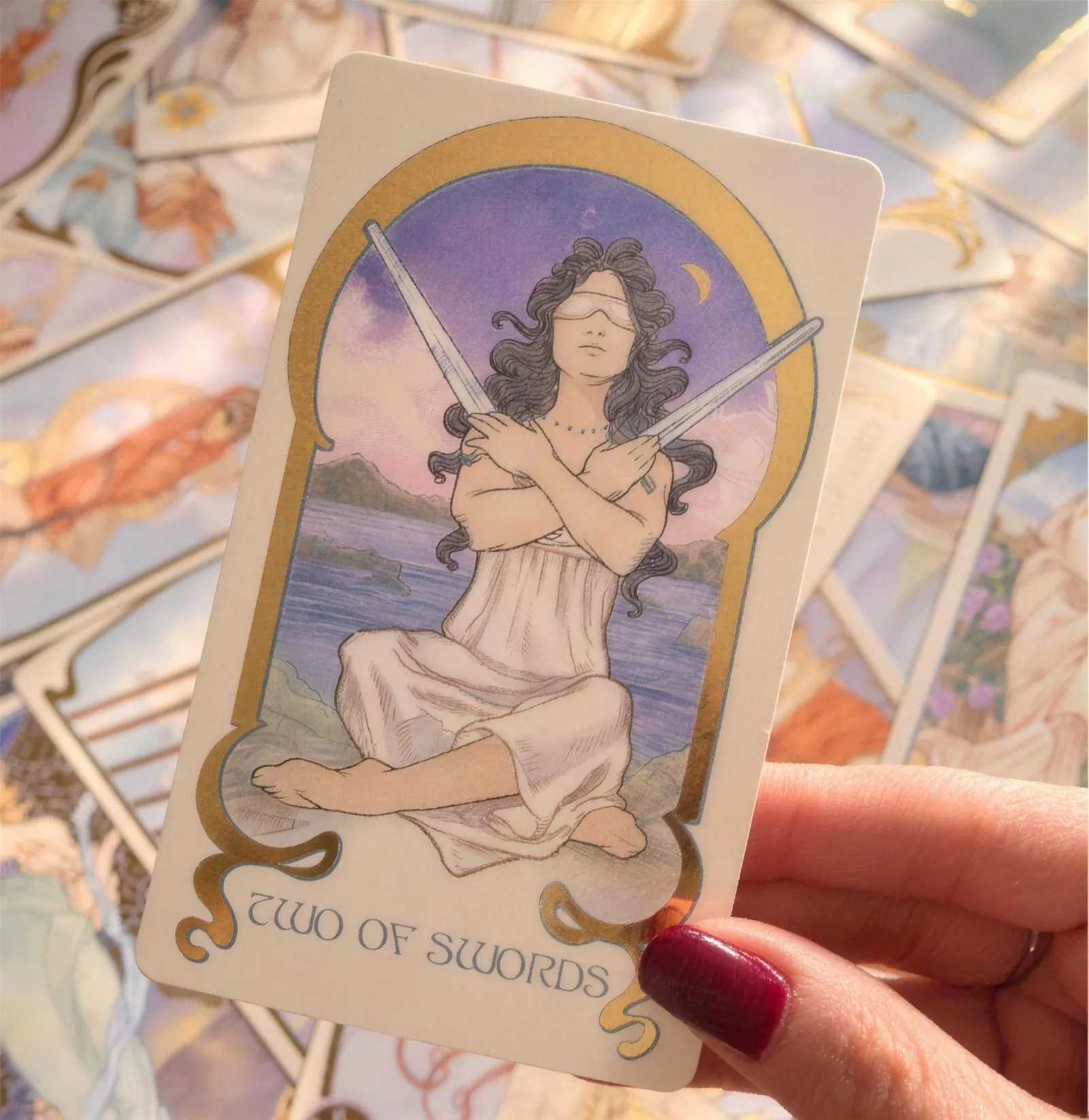 Mão segurando a carta Dois de Espadas sobre outras cartas de tarô espalhadas.