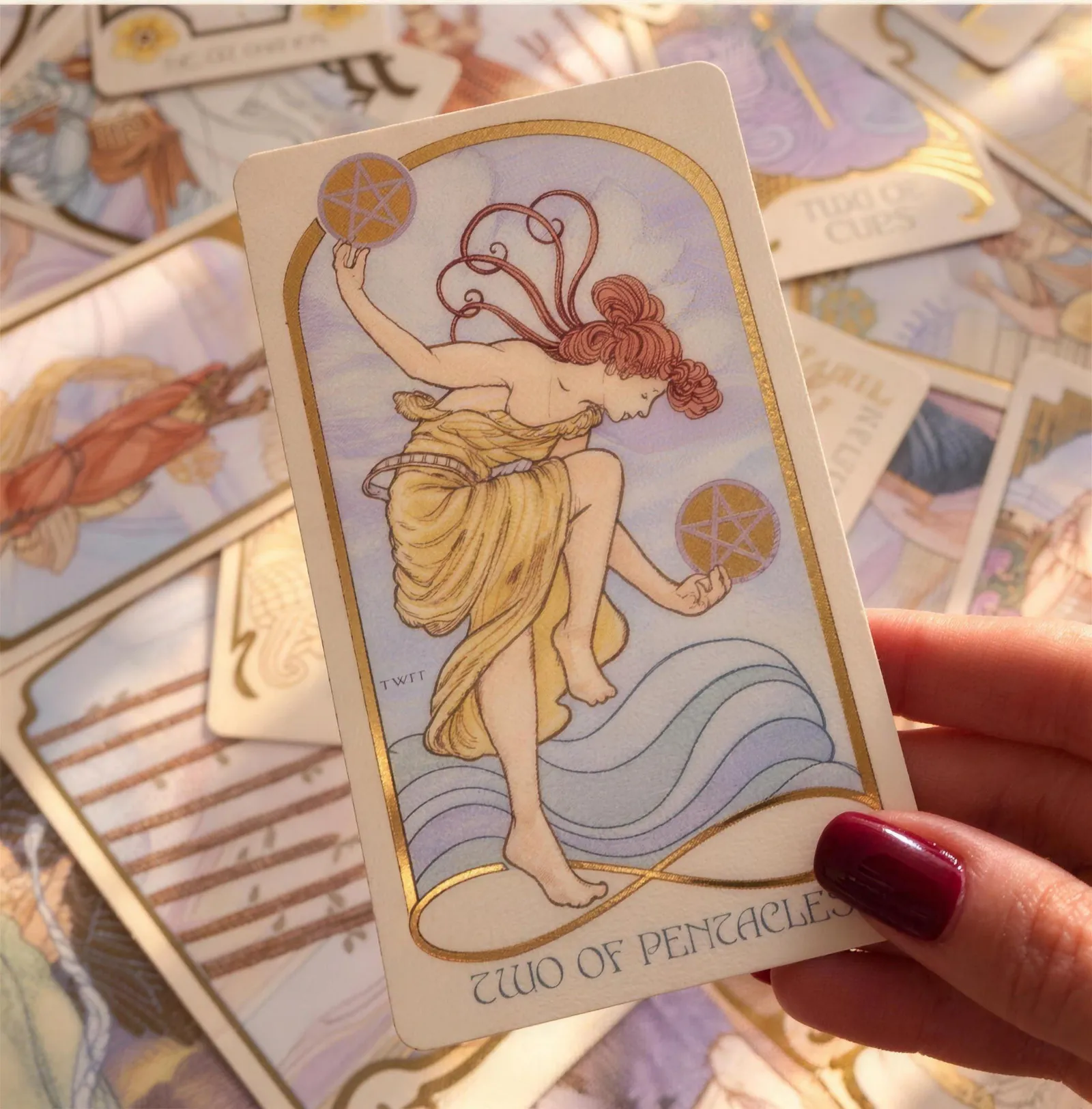 Mão segurando a carta Dois de Ouros sobre outras cartas de tarô espalhadas.