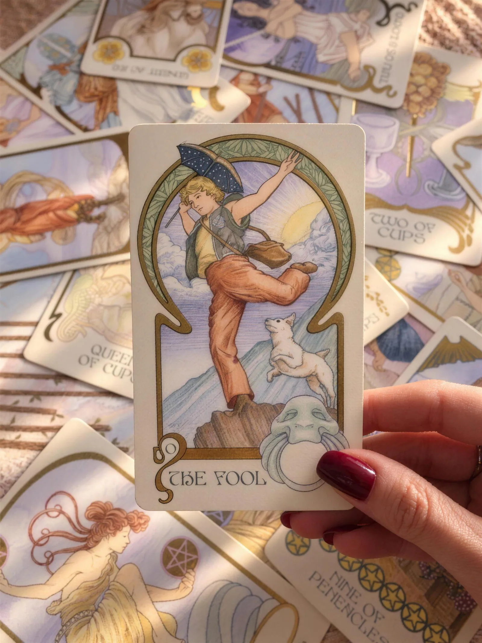 Mão segurando a carta O Louco, de um baralho de tarô com bordas douradas, sobre outras cartas espalhadas.